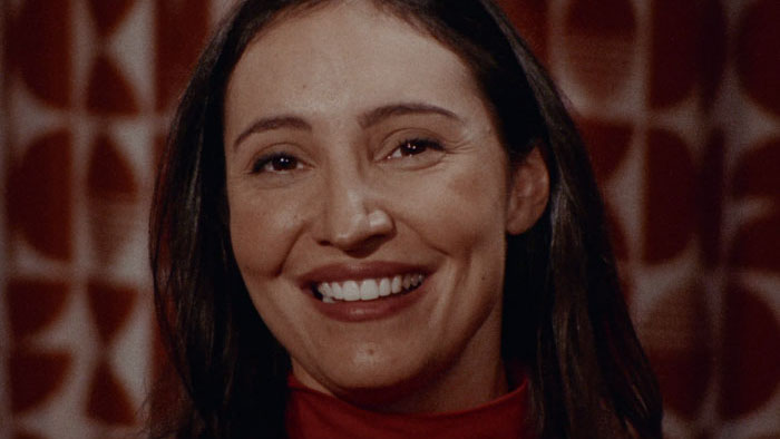 Close-up of a smiling woman in a red turtleneck with a retro patterned backdrop, from the film Neuro.