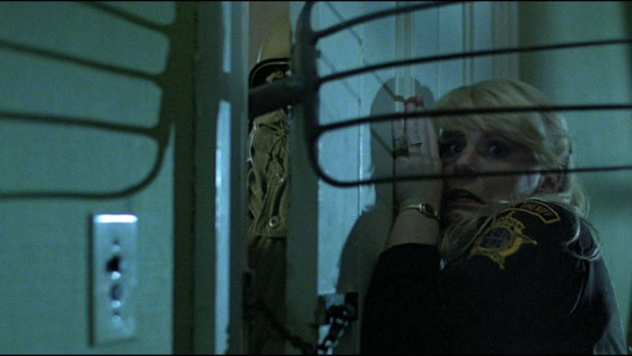 Terrified woman in a sheriff’s uniform leans against a door as a masked killer in military gear lurks outside in The Prowler (1981).