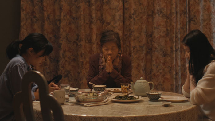 Andrea Bang, Cici Lau, and Grace Shen portray three generations of Asian women bowing their heads in prayer at a dinner table in a still from the film Surrender.