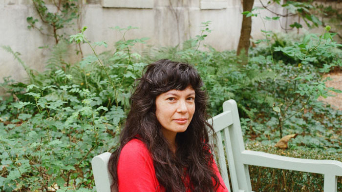Filmmaker Angeline Gragasin seated on a bench in a lush garden, wearing a red shirt, photographed in Paris in 2024