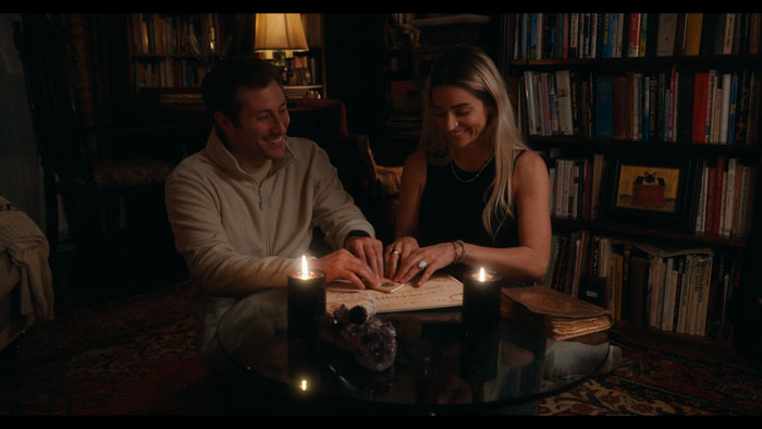 Cass and Tate use a spirit board by candlelight in a library room