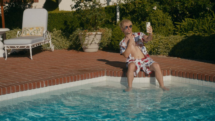 Man in floral shirt taking a selfie while lounging poolside with his feet in the water on a sunny day.