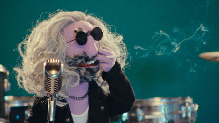 Purple muppet rockstar with long gray hair and sunglasses smokes a cigarette in front of a vintage microphone and drum set.