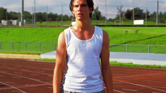 Brad Gorton as John Dixon stands on a high school running track in Get Real (1998), wearing athletic gear and looking conflicted.