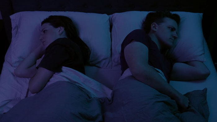 Brandon and Emma lie back-to-back in bed, emotionally distant