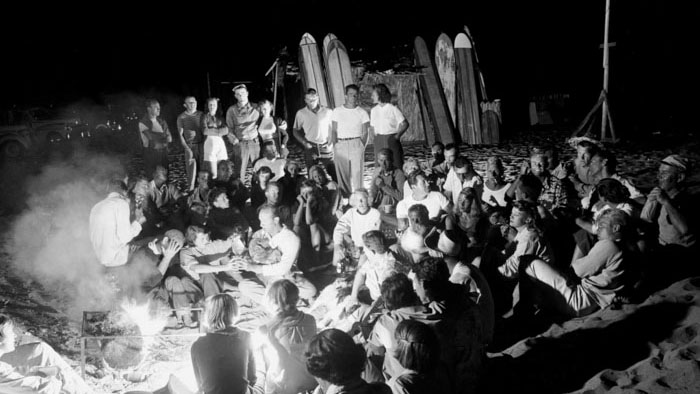 1950s Kookie beach luau with surfers gathered around bonfire at night