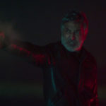 Pablo fires a gun in the dark, lit by red and green neon, in The Clouds (Las Nubes).