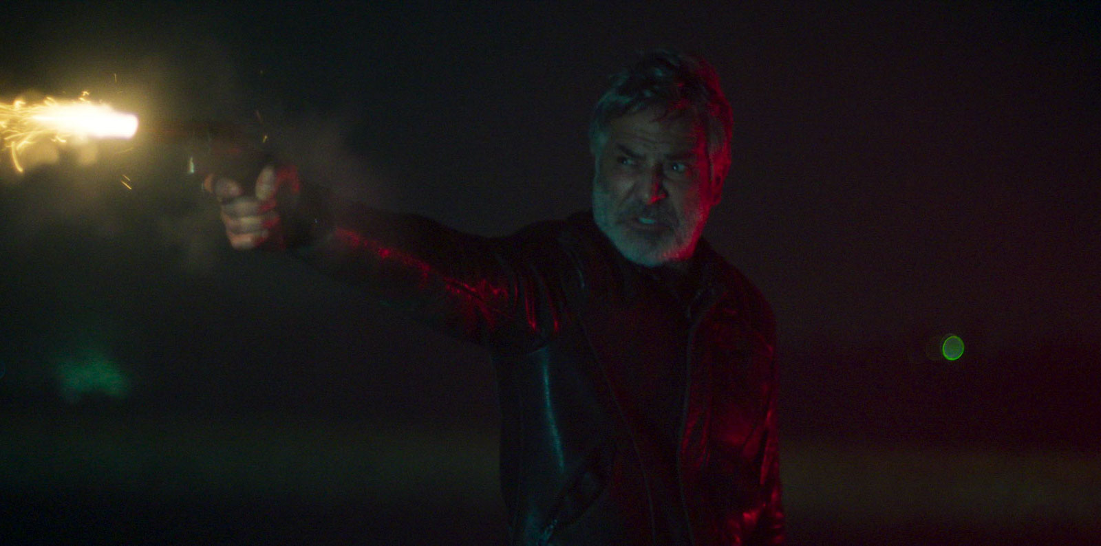 Pablo fires a gun in the dark, lit by red and green neon, in The Clouds (Las Nubes).
