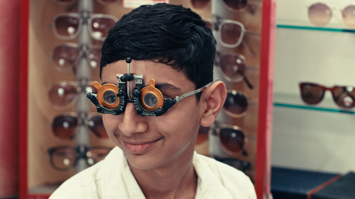 A young Indian boy smiles while wearing optometry test lenses inside a glasses store.