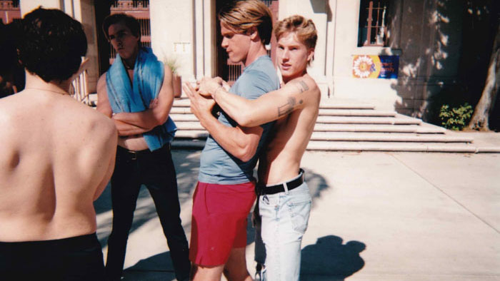 Two shirtless boys embrace in a tense moment on school grounds in Mean Boys