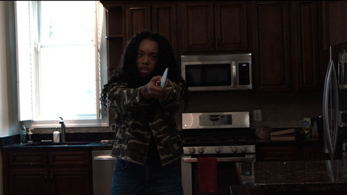 A woman in a camouflage jacket points a knife in a tense kitchen scene from The Final Podcast.
