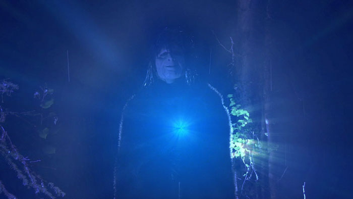 Black Turtle stands in the forest holding a glowing blue light in Alaskan Murder Mystery.