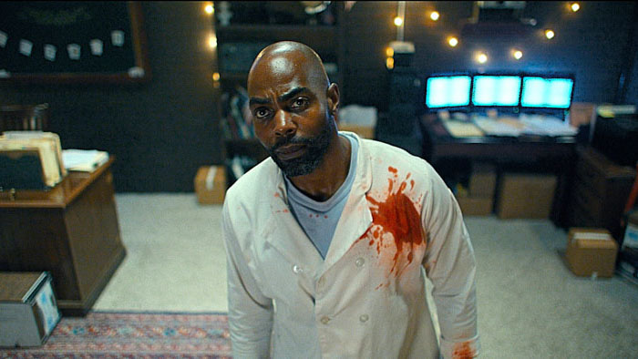 Clifton Duncan as Eric stands in the safe room wearing a blood-stained chef’s coat in American Deadbolt.