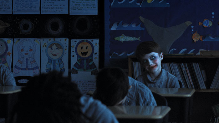 A child with smeared makeup smiles eerily in a dark classroom while other students have their heads down.