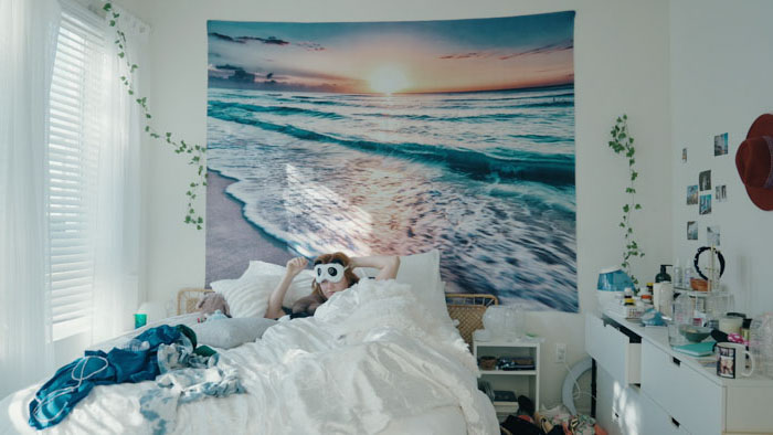 Woman lying in bed with owl eye mask under a beach sunset wall tapestry.