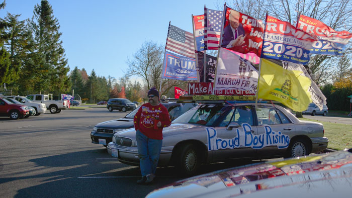 Man stands beside a car covered in Trump and political flags with “Proud Boy Rising” painted on the side.