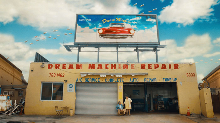 Marco and Isabela outside Dream Machine Repair auto shop beneath a billboard advertising flying cars.