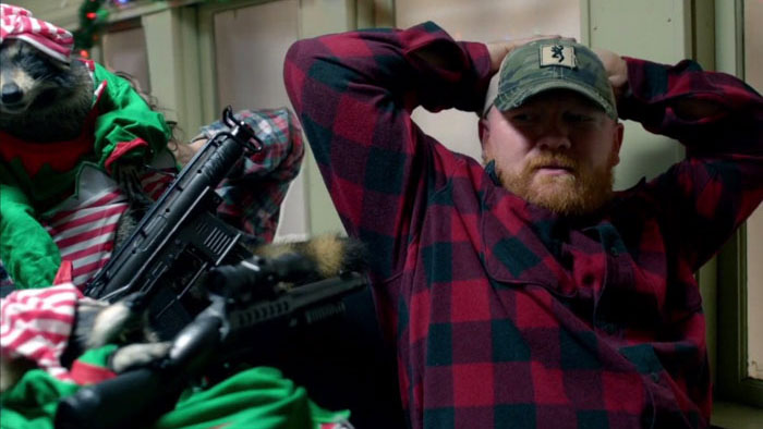 A man in a red plaid shirt held hostage by a raccoon dressed as an elf and armed with a rifle in Killer Raccoons 2: Dark Christmas in the Dark.