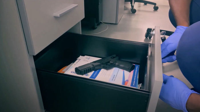 A gloved hand opens a desk drawer revealing a handgun inside a medical office.