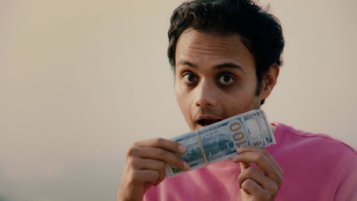 Ushmey Chakraborty as Ravi holding a stack of hundred-dollar bills in Gay, Asian, Immigrant