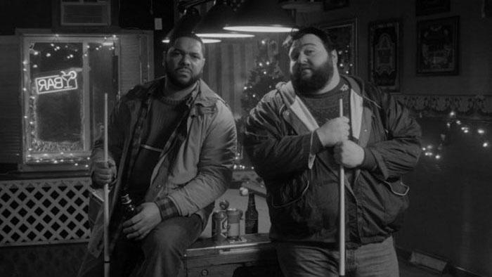 Two men holding pool cues sit by a bar table in a black-and-white scene from Stationed At Home. 