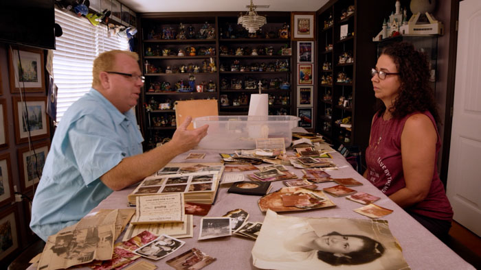 Michael Lester and Angela Lester sit at a table covered in family photos and documents in After the Devil is Dead. 