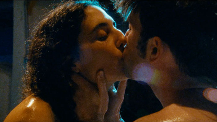 Thea and Ray share a passionate kiss under warm, shadowy lighting.