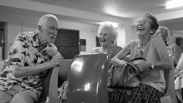 Black-and-white still from Flathead (2025) showing elderly residents laughing together in a modest community hall in Bundaberg, Australia.