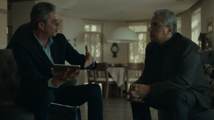 Two older men meet in a living room discussing a serious family matter in Happy Holidays (2025).