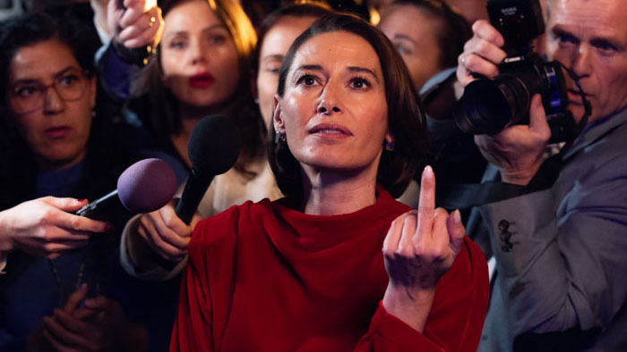 Prime Minister Alma Solvik in a red dress surrounded by reporters and microphones during a tense media scrum.