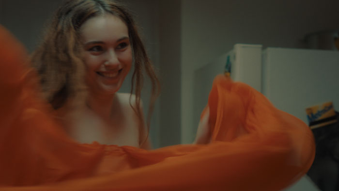 Masha (Ksena Samborska) smiles while holding a flowing orange dress in an apartment in Behind the Pose (2025).