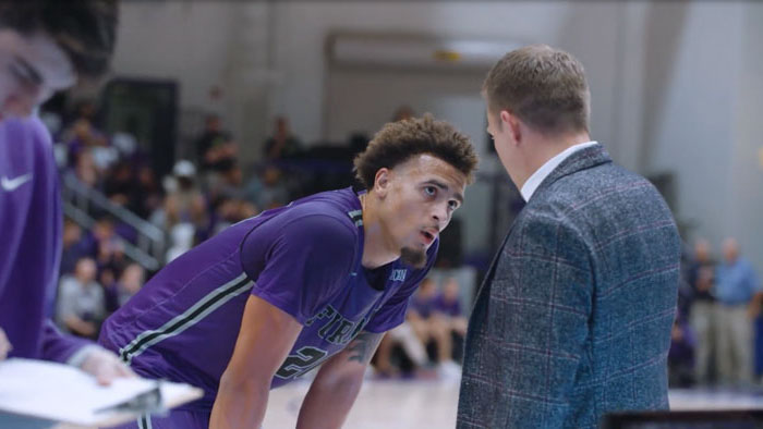 Furman basketball player in a purple jersey listens to coach during a game in Better Together: Furman’s Championship Quest.