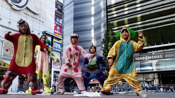Youths in colorful animal onesies dance in the street near TOHO CINEMAS in Kabukicho, Tokyo in Burn (2026).