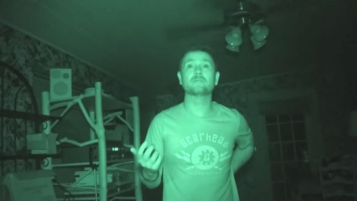 A man stands in a dim room filmed in green night vision, captured in found-footage style. 