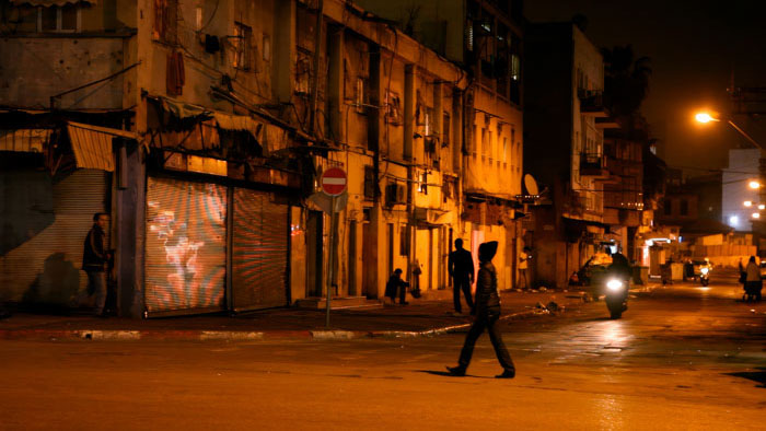 A dimly lit Tel Aviv street at night with pedestrians and buildings along the sidewalk.