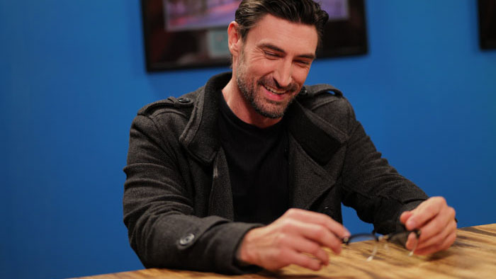 Ty Smith (John Wells) smiles at a table while holding his glasses in Learning You.