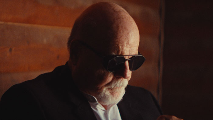 Michael Ironside as Dr. Jonathan Myers sits inside a cabin wearing dark sunglasses in warm, low light.