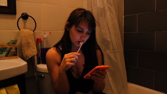 Marissa Peterson (Allison Sonson) sits in a bathroom brushing her teeth while looking at her phone in My Submission (2026).