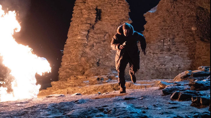 Jason Statham as Michael Mason runs through snowy ruins as flames explode nearby in Shelter (2026).