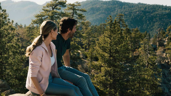 A couple sits on a rock ledge overlooking pine-covered mountains in daylight