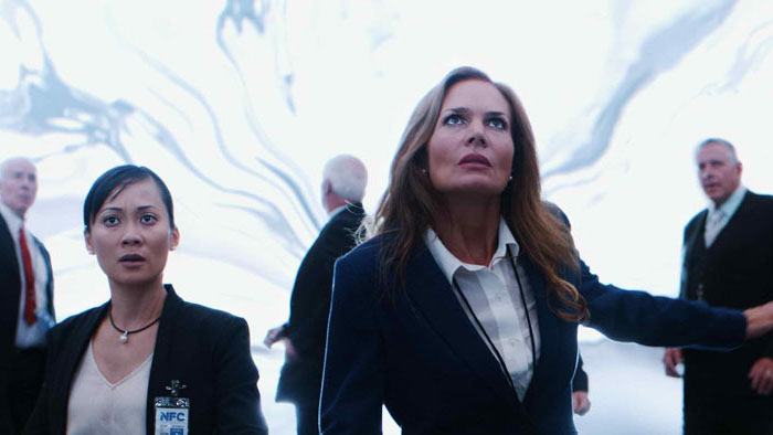 Two women with NFC badges stare upward as a bright portal-like phenomenon opens behind them in Space/Time (2026). 