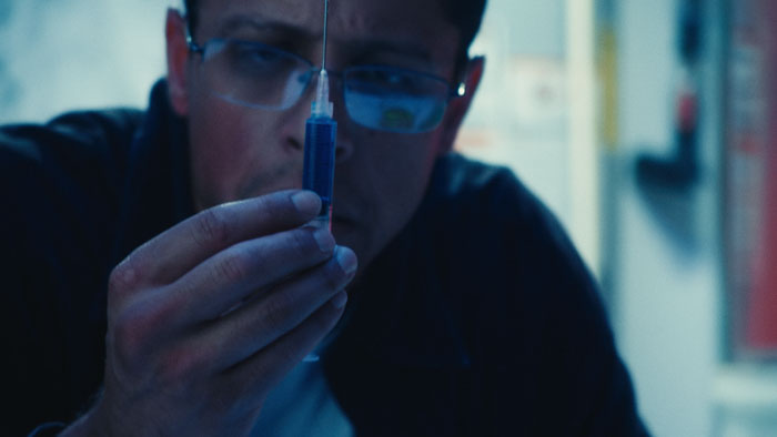 Richard holds a syringe of blue serum in The Geneticist (2026).