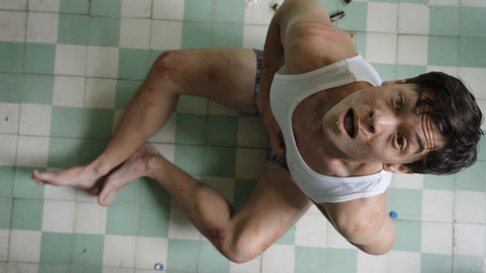 Will (Nate Nagvajara) sits on a tiled bathroom floor looking up, bruised and scraped, in The Itch (2025).