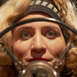 Close-up in The Soundman (2025) of a woman framed by a radio microphone and pop filter under warm studio lights.