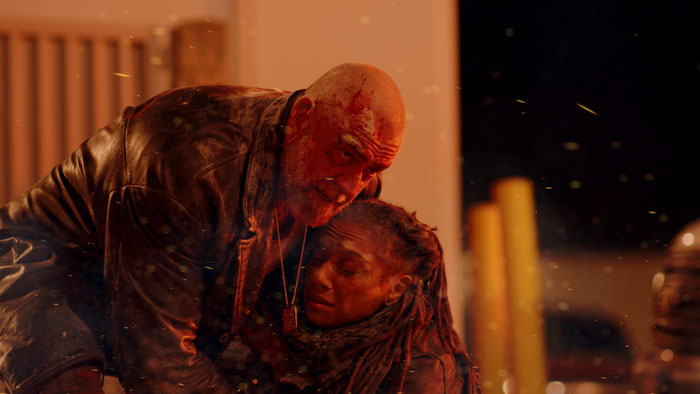 Bloodied man cradles an injured woman as embers drift in the air in Blackout (2026).