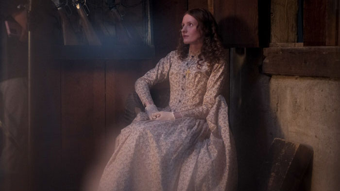 A woman in a long patterned dress sits indoors in soft light, looking off to the side.