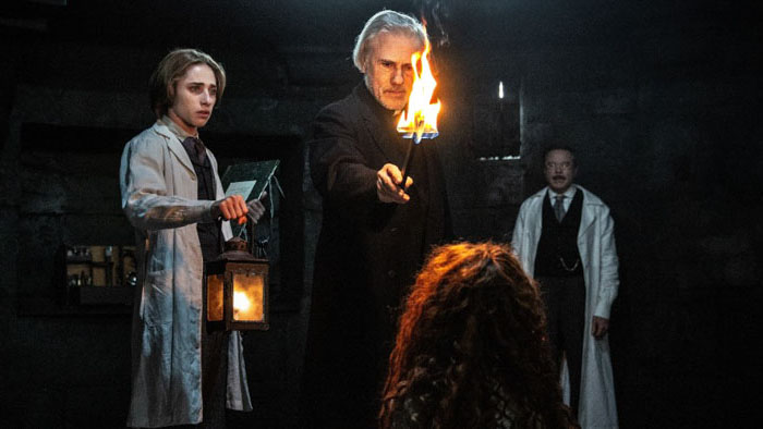 Three men confront a figure on the floor in a dark room, one holding a flaming torch and another holding a lantern in Dracula (2026). 