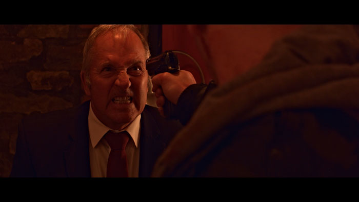 A man in a suit snarls as a gun is pressed to his head in a tense confrontation in Gifted Pain (2026)