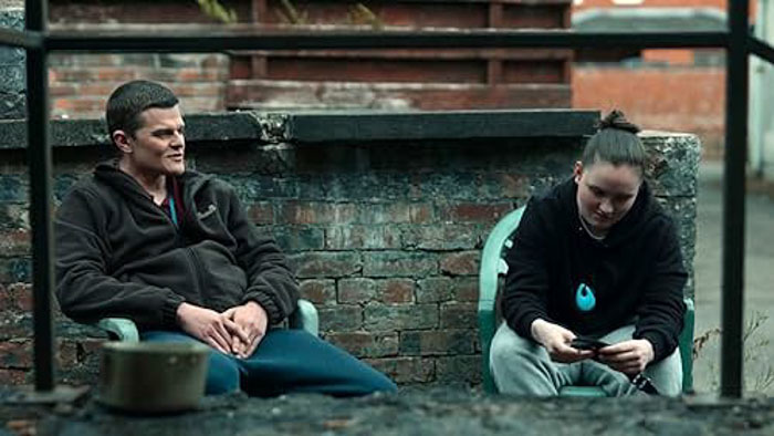 John sits outside with a woman in a brick courtyard in I Swear (2025).