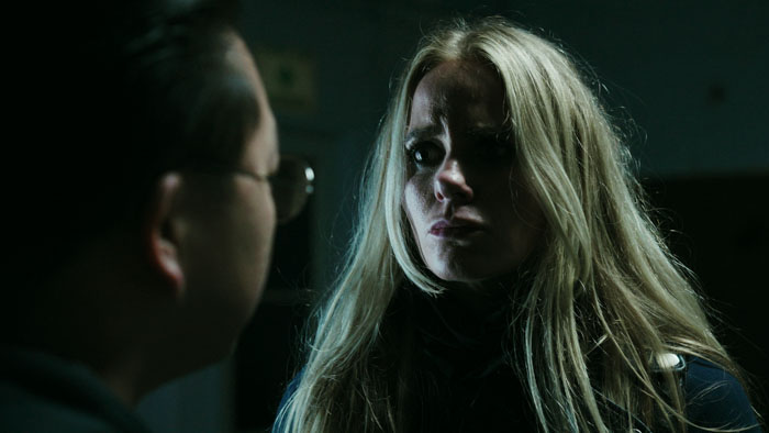 Tense close-up of a blonde woman confronting a man in a dimly lit scene from Last Hit (2026). 